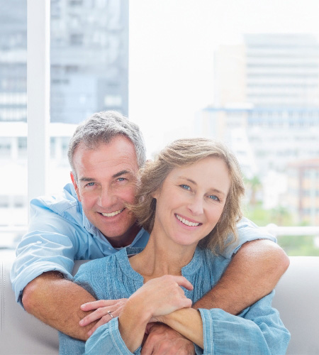 Older middle-aged man with gray hair standing behind a couch and hugging his wife who is sitting on it